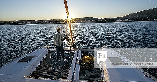 Junger Mann steht am Vorsegel  Sonnenuntergang mit Sonnenstern  Beim Segeln mit einem Segel-Katamaran  Dodekanes  Griechenland  Europa