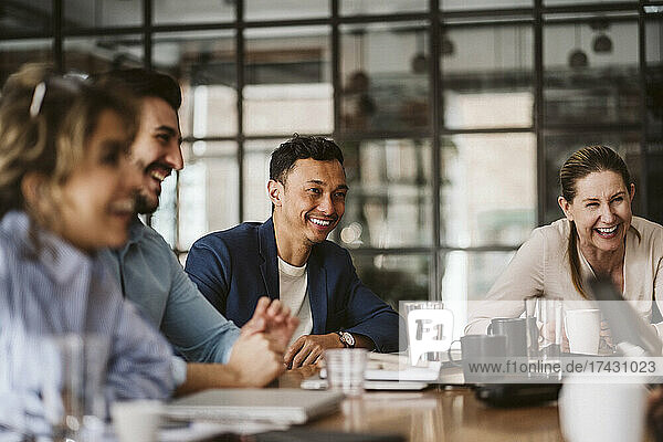 Smiling businessman looking away while sitting amidst cheerful colleagues at conference table