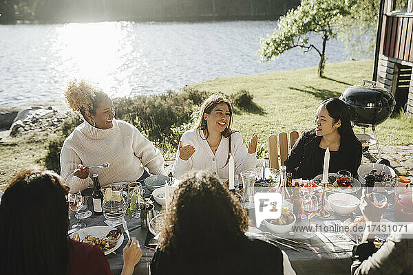 Glückliche Frau im Gespräch mit weiblichen Freunden  während sie eine Dinnerparty im Hinterhof an einem sonnigen Tag genießt
