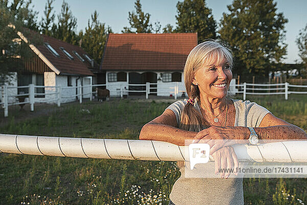 Lächelnde ältere Landarbeiterin lehnt am Zaun eines Bauernhofs