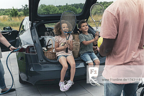 Geschwister halten Tennisschläger  während sie im Kofferraum sitzen und ihren Vater ansehen
