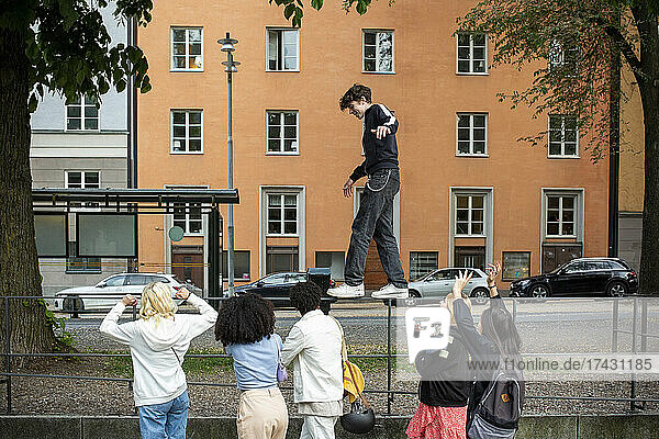 Männlicher Teenager balanciert auf einem Geländer  während Freunde auf dem Fußweg jubeln