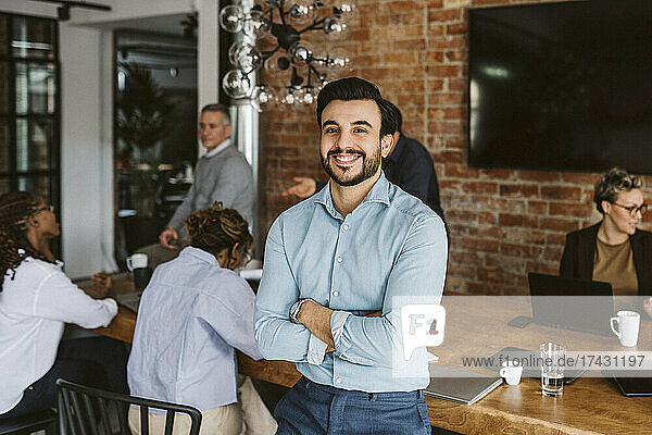 Portrait of smiling male entrepreneur standing with arms crossed while colleagues discussing in background