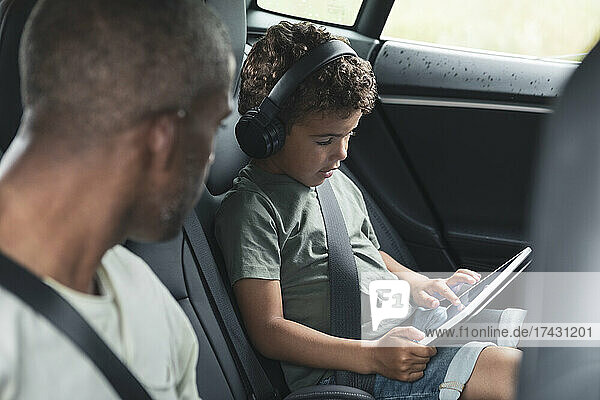 Mann schaut seinem Sohn zu  der im Elektroauto ein Video auf einem digitalen Tablet anschaut