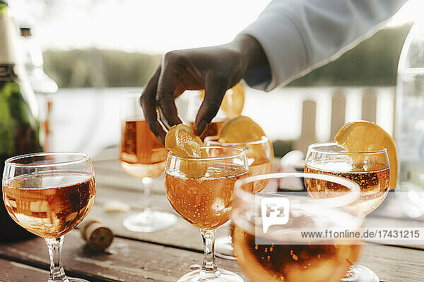 Abgeschnittene Hand einer Frau  die eine Orangenscheibe auf einem Weinglas am Tisch hält