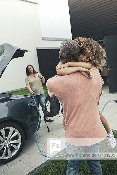 Rückansicht eines Mannes  der seine Tochter trägt  während er neben einem Elektroauto steht