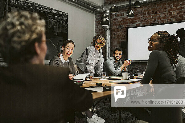 Smiling male and female professionals looking at mature entrepreneur while sitting at conference table