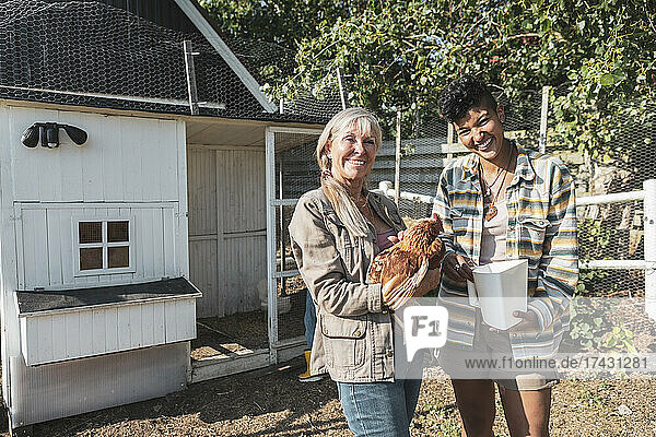 Porträt einer lächelnden gemischtrassigen Mutter und Tochter mit Huhn auf einem Bauernhof an einem sonnigen Tag