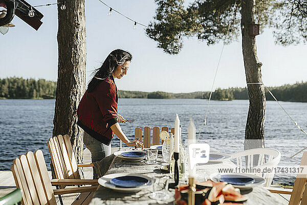 Young woman setting table by lake on sunny day
