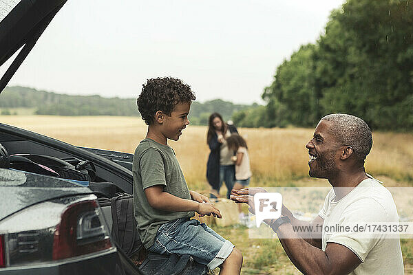 Lächelnder Mann spielt mit seinem Sohn  der im Kofferraum eines Autos sitzt  während eines Picknicks