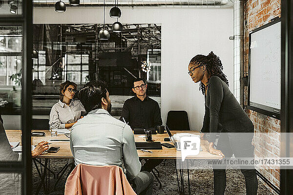 Geschäftsfrau lehnt sich an einen Konferenztisch  während sie mit Kollegen im Sitzungssaal diskutiert