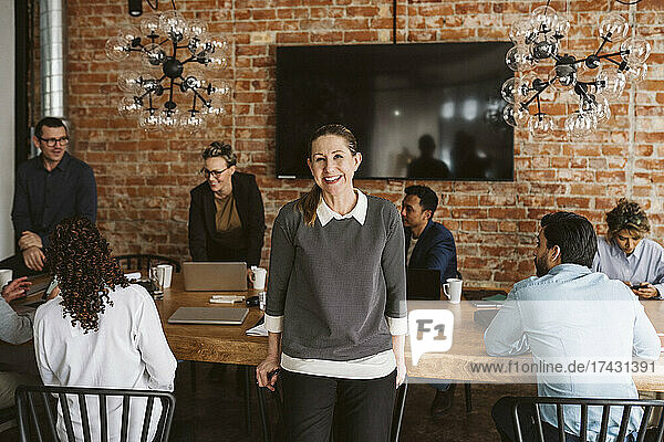 Porträt einer glücklichen reifen Geschäftsfrau im Stehen  während Kollegen im Hintergrund diskutieren
