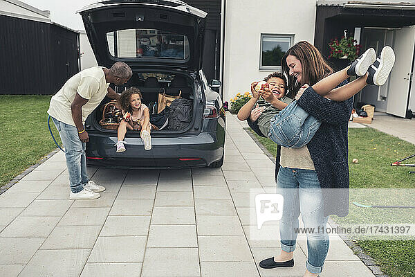 Lächelnde Frau  die ihren Sohn trägt  während der Mann seine im Kofferraum sitzende Tochter anschaut