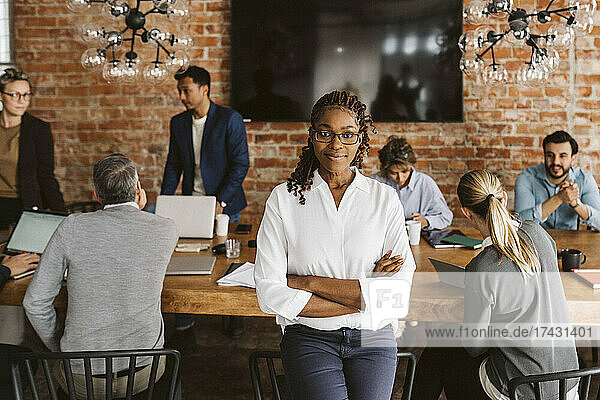 Portrait of confident female entrepreneur standing with arms crossed while colleagues discussing in background