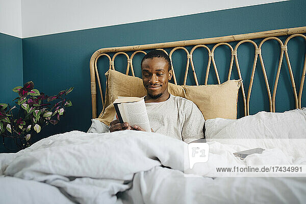 Lächelnder Mann im mittleren Erwachsenenalter  der zu Hause im Bett ein Buch liest