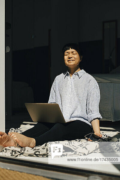 Lächelnde Frau mit Laptop  die zu Hause im Sonnenlicht sitzt