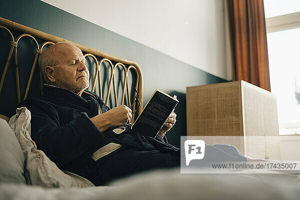 Älterer Mann mit geschlossenen Augen hält ein Buch  während er auf dem Bett sitzt