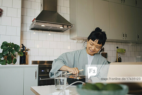 Lächelnde Frau beim Öffnen eines Lebensmittelbehälters am Tisch in der Küche