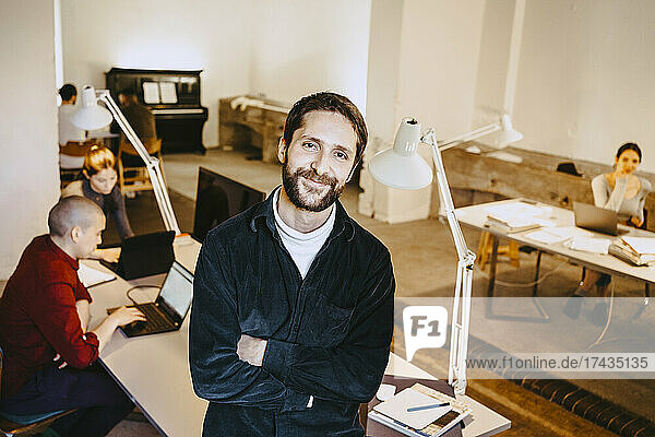 Portrait of smiling male programmer standing with arms crossed while colleagues working at desk in office