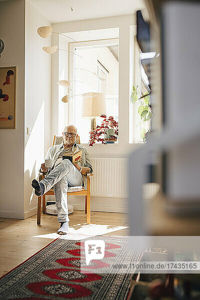 Älterer Mann liest ein Buch,  während er zu Hause am Fenster Kaffee trinkt