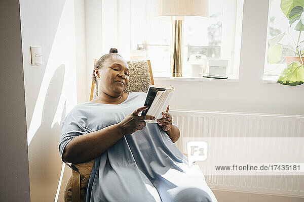 Lächelnde Frau liest ein Buch und sitzt auf einem Stuhl im Wohnzimmer