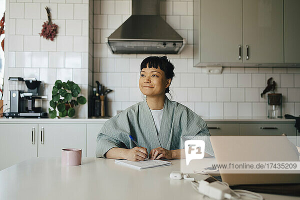 Smiling woman looking away while writing in book over kitchen island at home