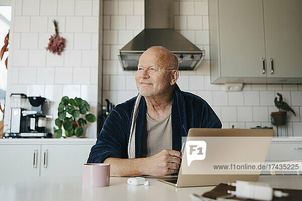 Smiling senior man with laptop looking away while sitting in kitchen at home