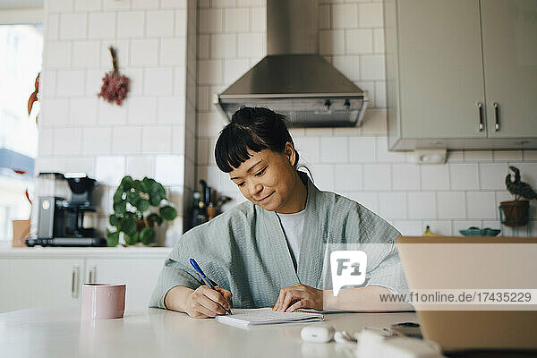 Young woman in bathrobe writing in book at domestic kitchen