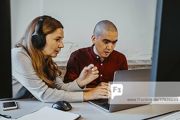 Mature female hacker discussing with male colleague over laptop at creative office