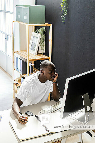 Male freelancer sitting with spiral notebook looking at desktop computer in studio