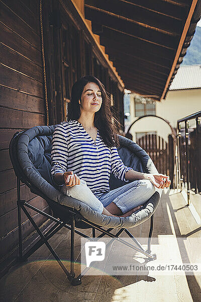 Frau meditiert  während sie mit gekreuzten Beinen auf einem Stuhl auf dem Balkon sitzt