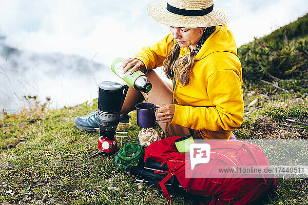 Woman wearing hat pouring tea in cup while sitting by backpack on mountain