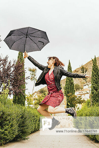 Happy mid adult woman jumping with umbrella during rain at park