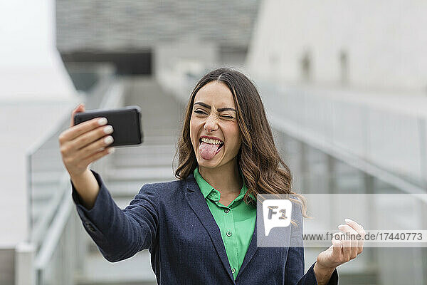 Cheerful businesswoman sticking out tongue while taking selfie through smart phone