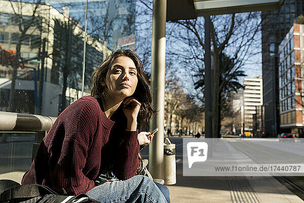 Young woman waiting for tram while sitting on bench in city