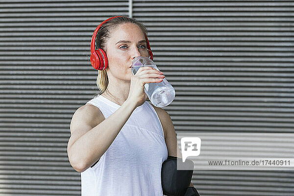 Mittlere erwachsene Sportlerin trägt Kopfhörer und trinkt Wasser an der Wand