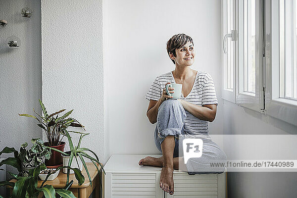 Lächelnde Frau mittleren Alters mit Tasse  die durch das Fenster schaut  während sie zu Hause sitzt