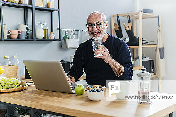 Lächelnder Mann mit Laptop hält Trinkglas am Küchentisch