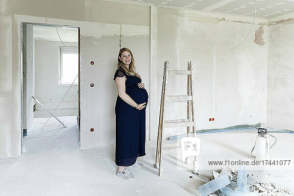 Smiling pregnant woman touching abdomen at house under renovation