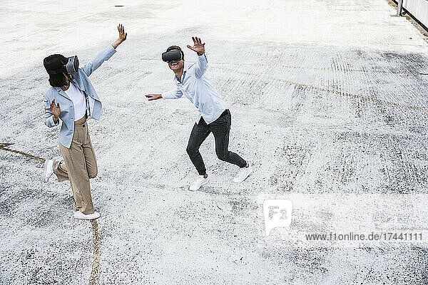 Playful male and female business colleagues with arms outstretched using virtual reality headset on rooftop