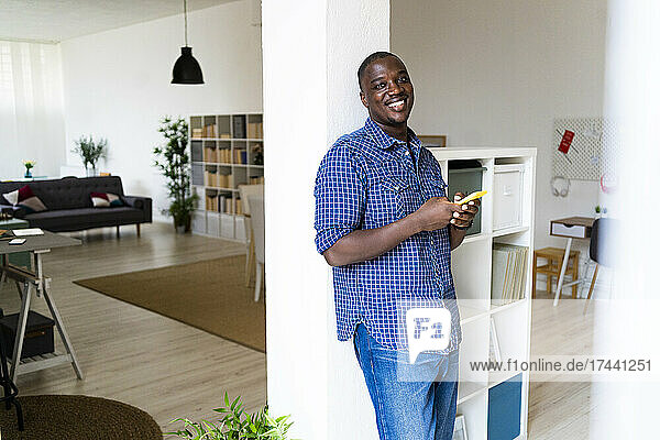 Smiling man with mobile phone leaning on column at home