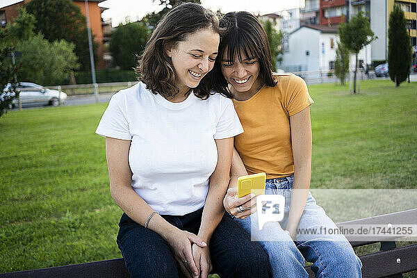Happy lesbian couple using smart phone on bench