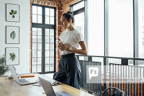 Female business professional with coffee cup looking at laptop while standing in office