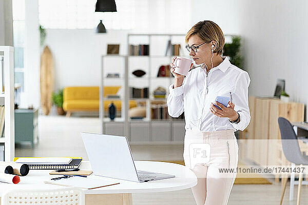 Businesswoman with mobile phone drinking coffee in office