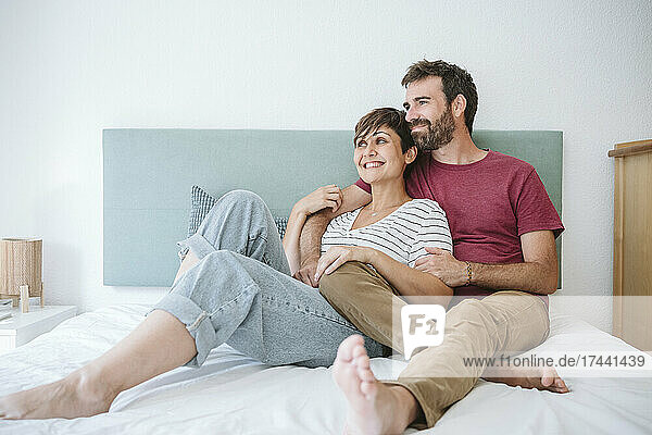 Glückliches Paar mittleren Alters schaut weg  während es zu Hause zusammen im Bett sitzt