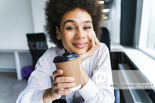 Smiling businesswoman with hand on chin holding disposable cup in office
