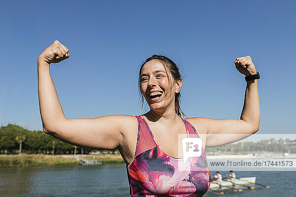 Sportswoman laughing while flexing muscles at riverbank
