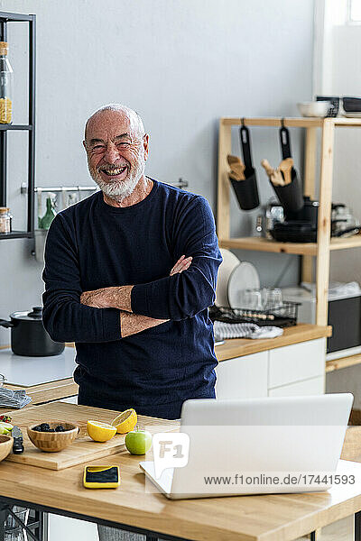 Cheerful senior man with arms crossed standing in kitchen at home