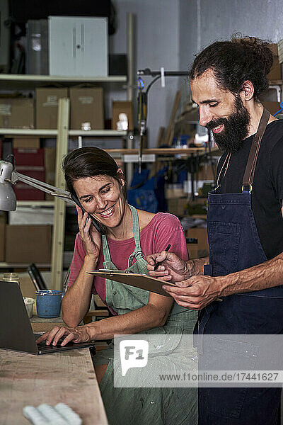 Happy female entrepreneur talking on smart phone while sitting by man holding clipboard in workshop
