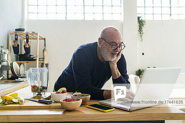 Senior man using laptop while leaning on kitchen counter